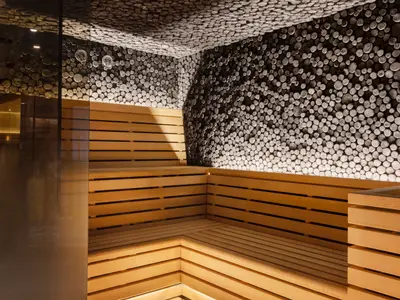 Innenansicht einer stilvollen Sauna an Bord der Norwegian Viva. Mehrstufige Holzbänke und Wände, die mit unzähligen runden Holzscheiben verziert sind. Warmes, indirektes Licht beleuchtet den Raum, der durch eine Glastür begrenzt wird. Zwei Thermometer sind an der Wand zu sehen.