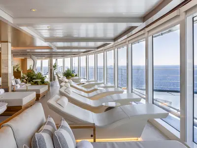 Die Thermal Lounge an Bord der Norwegian Viva bietet eine Reihe von eleganten, weissen Liegestühlen mit Blick auf das blaue Meer durch grosse Fenster. Üppige Pflanzen und warme Holzelemente schaffen eine ruhige Atmosphäre.