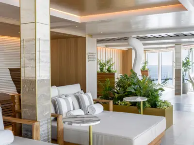 Die Thermal Lounge an Bord der Norwegian Viva zeigt bequeme Daybeds mit Kissen und Handtüchern, umgeben von üppigen Pflanzen und modernen Marmorsäulen. Große Fensterfronten bieten einen weiten Blick auf das offene Meer. Die helle, holzvertäfelte Atmosphäre lädt zum Entspannen ein.