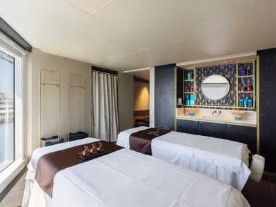 Luxuriöser Doppel-Behandlungsraum im Mandara Spa der Norwegian Viva. Zwei Massagebetten, Waschtisch mit zwei Waschbecken, Spiegel, Pflegeprodukten und große Fenster mit Ausblick sind zu sehen.