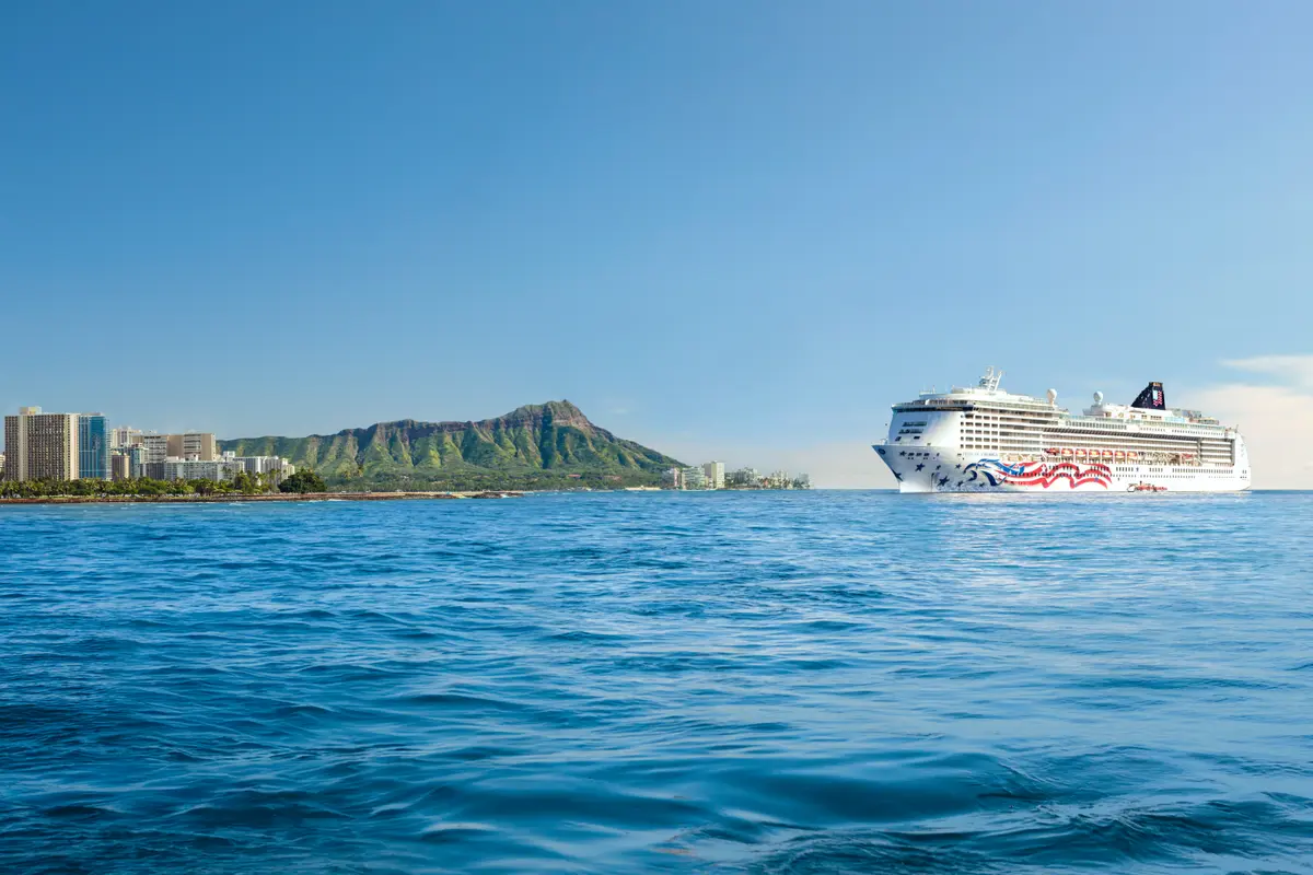 Die Pride of America fährt auf blauem Meer, im Hintergrund die Skyline von Honolulu und Diamond Head auf Oʻahu, Hawaii. Heller Himmel.