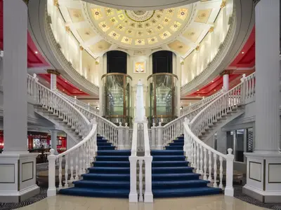 Das Capitol Atrium an Bord der Pride of America. Eine breite Doppeltreppe mit blauem Teppich führt zu gläsernen Aufzügen und einem Obelisken unter einer beleuchteten Kuppel.