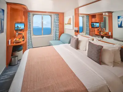 Helle Aussenkabine an Bord der Pride of America mit Blick auf das Meer und Inseln. Zu sehen sind ein großes Bett, ein Sofa und ein Schreibtisch mit TV, Obst und Champagner.