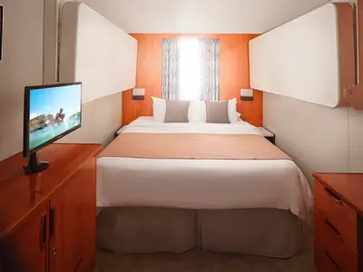 Moderne Familienkabine an Bord der Pride of America. Zentral ein Doppelbett mit weißer Bettwäsche und orangen Akzentkissen. Orange Wand mit Fenster im Hintergrund. Links ein Holzschrank mit Fernseher.