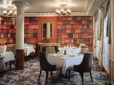 Ein stilvolles Restaurant an Bord der Pride of America. Der Raum ist mit eleganten Tischen, Stühlen und einer Wanddekoration im Stil einer Bibliothek ausgestattet. Viele Weinflaschen und Gläser stehen bereit.