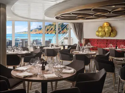 Ein elegantes Restaurant an Bord der Pride of America. Gedeckte Tische und moderne Stuehle. Grosse Fenster zeigen Meer und gruene Berge. Goldene Wandkunst.