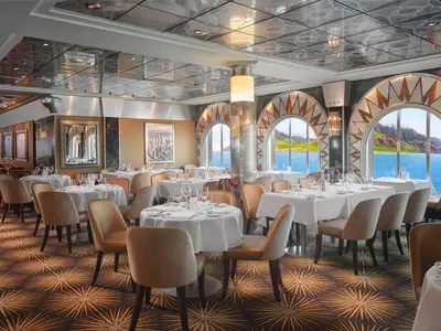 Blick in das stilvolle Skyline Restaurant an Bord der Pride of America. Viele Tische sind gedeckt, dahinter bieten große Bogenfenster einen beeindruckenden Meer- und Küstenblick mit grünen Bergen.