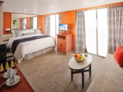Blick in eine geräumige Kabine der Pride of America: Grosses Bett mit blauer Tagesdecke, dahinter Spiegel. Rechts ein Fernseher, ein Couchtisch mit Obst und Champagner, sowie ein Sofa vor grossen Fenstern mit gemusterten Vorhängen.