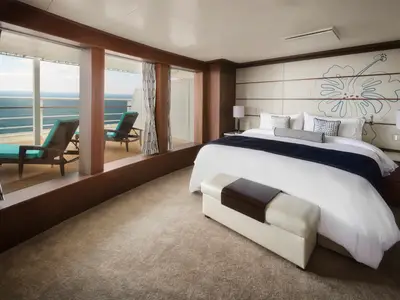 Ein luxuriöses Schlafzimmer in einer Suite der Pride of America. Es zeigt ein großes Bett, moderne Möbel und einen Balkon mit Meerblick. Zwei blaue Sonnenliegen stehen auf dem Deck.