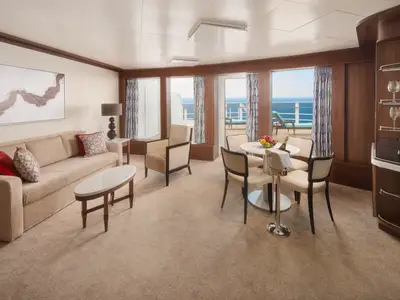 Ein luxuriöser Wohnbereich an Bord der Pride of America. Beige Möbel, ein Esstisch mit frischem Obst und ein Balkon mit Meerblick sind zu sehen.
