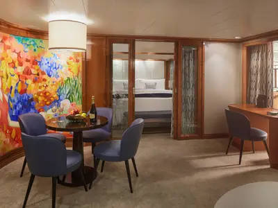 Die luxuriöse Owner's Suite an Bord der Pride of America zeigt einen Wohnbereich mit Esstisch, Kunstarbeit und einen separaten Schlafbereich. Rechts ein Schreibtisch mit Meerblick.