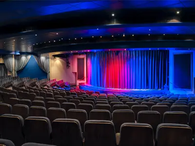 Das leere Hollywood Theater an Bord der Pride of America zeigt eine Bühne mit rot und blau beleuchteten Vorhängen. Davor reihen sich viele dunkelblaue Sitze in einem eleganten Ambiente.