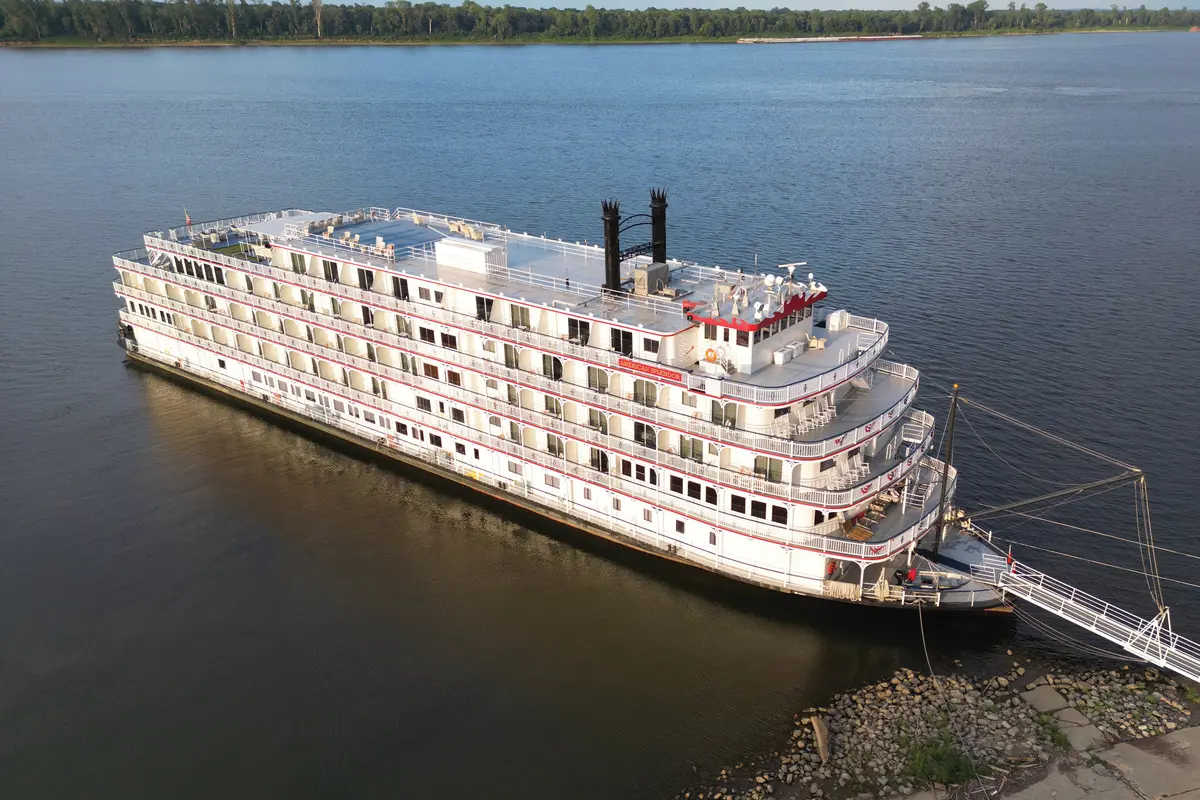 Luftaufnahme eines großen, weißen Flusskreuzfahrtschiffs namens "American Splendor" mit mehreren Decks, roten Akzenten und schwarzen Schornsteinen, ruhig auf blauem Wasser liegend.