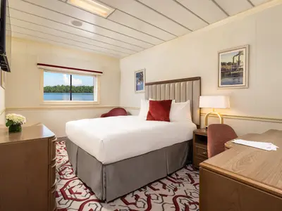 Eine helle Kabine an Bord der American Splendor. Zu sehen sind ein Bett, ein Schreibtisch, TV und ein Fenster mit Blick auf einen Fluss. Die Einrichtung ist modern mit roten Akzenten.