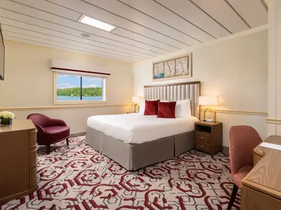 Eine Kabine des Flusskreuzfahrtschiffs American Splendor mit Doppelbett, Sessel, Schreibtisch. Fensterblick auf bewaldete Flusslandschaft.