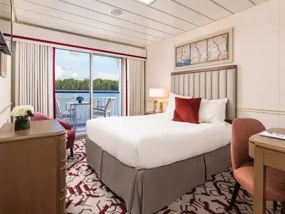 Geräumige Kabine des Flusskreuzfahrtschiffs American Splendor mit Doppelbett, Schreibtisch und möbliertem Balkon. Flussblick auf grüne Ufer.
