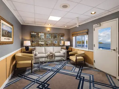 Eine helle Lounge auf der American Splendor mit gemütlichen Sitzmöbeln, geometrischem Teppich und Fenstern zur Flusslandschaft. Die graue Wand ist mit Spiegeln dekoriert.
