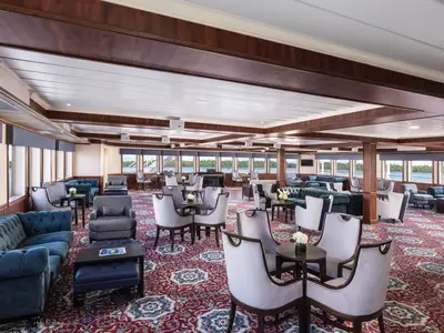 Lounge auf dem Flusskreuzfahrtschiff American Splendor. Blaue Sofas, graue Sessel, helle Stühle, rote Teppiche. Große Fenster zeigen die Flusslandschaft.