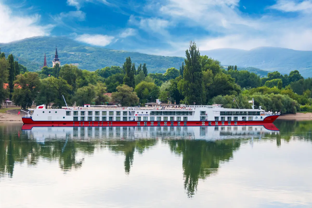 Flusskreuzfahrtschiff MS Belvedere (Nicko Cruises) auf dem Wasser. Rot-weißer Rumpf spiegelt sich. Im Hintergrund: bewaldete Hügel, Kirchtürme, blauer Himmel.