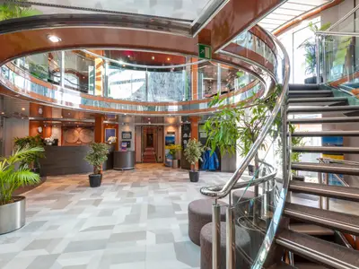 Blick in die helle Lobby des Flusskreuzfahrtschiffs Belvedere mit einer eleganten Wendeltreppe und Glasbalustrade. Im Hintergrund befinden sich der Empfangstresen und eine Weltkarte. Zahlreiche Grünpflanzen schmücken den Raum.