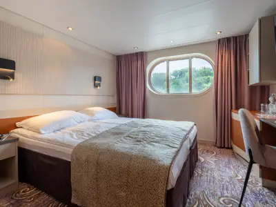 Kabine des Flusskreuzfahrtschiffs Belvedere: Doppelbett, ovales Fenster mit Ausblick auf grüne Landschaft, Schreibtisch mit Stuhl und montiertem Fernseher.