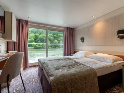 Kabine des Flusskreuzfahrtschiffs Belvedere mit Doppelbett, Schreibtisch und Glastür zu einem Balkon. Dahinter der Fluss und bewaldete Hügel. Helle und moderne Einrichtung.