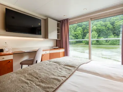 Helle Kabine auf der Belvedere mit Betten, Schreibtisch und TV. Große Fensterfront bietet Blick auf Fluss und bewaldete Ufer.