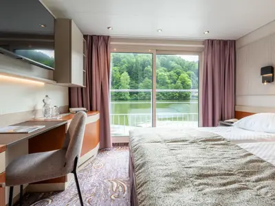 Helle Kabine des Flusskreuzfahrtschiffs Belvedere mit Schreibtisch, Bett und großem Panoramafenster. Der Blick geht auf eine grüne, bewaldete Flusslandschaft und das Wasser. Eine Wasserflasche steht auf dem Tisch.