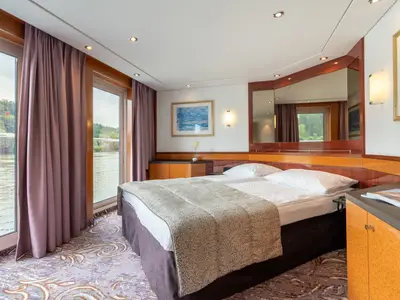 Innenansicht einer modernen Doppelkabine an Bord der Belvedere. Sie zeigt ein komfortables Bett, große Panoramafenster mit Flussblick, Holzmöbel und einen farbigen Teppich.
