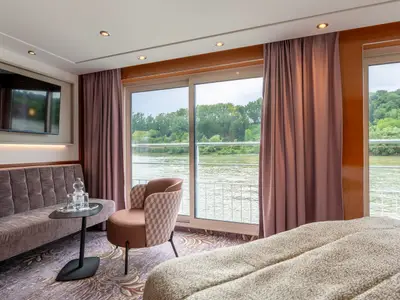Innenansicht einer geräumigen Kabine des Flusskreuzfahrtschiffs Belvedere von nicko cruises mit Sofa, Sessel und Blick auf den Fluss durch eine breite Glasfront.