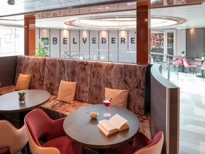 Moderne Bar-Lounge auf einem Kreuzfahrtschiff mit Sitzbänken, runden Holztischen und roten Stühlen. Ein Cocktail und ein Buch liegen auf einem Tisch. An der Wand steht der Schriftzug "BELVEDERE".