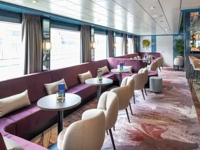 Moderne Lounge des Flusskreuzfahrtschiffs Belvedere. Man sieht lila Sofas an Fenstern, runde Tische, beige Sessel, eine Bar und floralen Teppich.