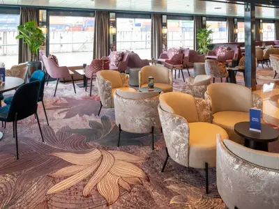 Eleganter Lounge-Bereich des Flusskreuzfahrtschiffs Belvedere. Modern eingerichteter Raum mit vielen Sitzgelegenheiten, floralem Teppich und Blick auf einen Hafen.