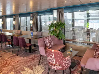 Die helle Lounge des Flusskreuzfahrtschiffs Belvedere. Das Bild zeigt Tische mit lila Stühlen, beigen Sitzbänken und großen Fenstern. Ein gemusterter Teppich ziert den Boden.