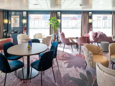 Die farbenfrohe Lounge des Flusskreuzfahrtschiffs Belvedere. Verschiedene Sitzgelegenheiten, floraler Teppich und Fenster mit Blick auf eine Hafenanlage. Die Belvedere lädt zum Verweilen ein.