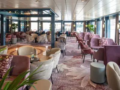 Die stilvolle Lounge des Flusskreuzfahrtschiffs Belvedere zeigt moderne Sitzgruppen in Violett und Beige. Große Panoramafenster bieten Ausblick, der Teppich ist floral gemustert.
