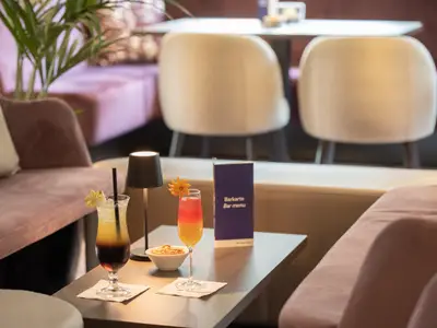 Eine Nahaufnahme zeigt zwei bunte Cocktails auf einem Holztisch in einer modernen Bar-Lounge. Im Hintergrund sind weiche, lilafarbene Sofas und Stühle zu sehen. Eine Barkarte und ein kleines Licht zieren den Tisch.