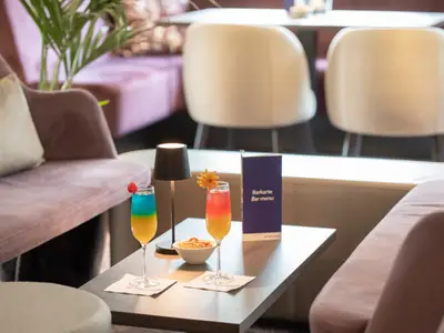 Zwei farbige Cocktails auf einem Tisch in einer modernen Bar-Lounge, umgeben von lilafarbenen Sofas und hellen Stühlen. Eine Lampe und die Barkarte sind sichtbar.