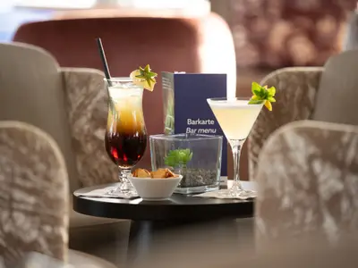 Zwei bunte Cocktails auf einem kleinen runden Tisch in einer stilvollen Bar-Lounge. Dazu stehen eine Schale mit Snacks, ein Dekoelement und eine Barkarte. Im Hintergrund sind beige Sessel und diffuse Beleuchtung zu sehen.