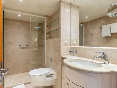 Modernes, helles Badezimmer einer Suite. Marmorfliesen, begehbare Dusche mit Haltegriff, WC, Waschtisch und Chromarmatur sind sichtbar.