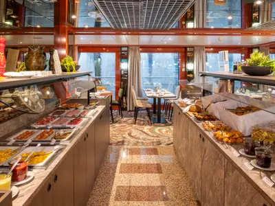 Der Buffetbereich des Flusskreuzfahrtschiffs Belvedere mit vielfältigen Speisen wie Brötchen, Aufschnitt, Eiern und Gebäck. Im Hintergrund sind Tische und große Fenster zu sehen.