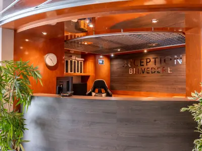 Rezeption des Flusskreuzfahrtschiffs Belvedere. Dunkle Theke, Arbeitsplatz, Stuhl. Orange Wand mit "RECEPTION BELVEDERE" auf Holz. Grüne Pflanzen.