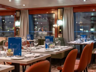 Elegantes Restaurant auf dem Flusskreuzfahrtschiff belvedere. Gedeckte Tische mit Gläsern und blauen Menükarten. Nachtblick auf beleuchtete Stadt durch Fenster. Warme Atmosphäre an Bord der belvedere.