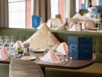 Gedeckter Tisch im Restaurant des Flusskreuzfahrtschiffs Belvedere mit Gläsern, Besteck, Servietten und nicko cruises Menükarte. Grüne Bänke im Hintergrund.