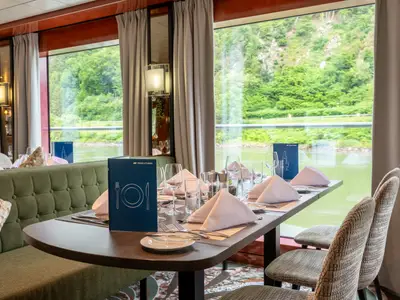 Ein Innenblick auf das Restaurant des Flusskreuzfahrtschiffs Belvedere. Die Tische sind gedeckt mit Besteck, Gläsern und rosa Servietten. Große Fenster bieten eine Aussicht auf eine grüne Flusslandschaft.