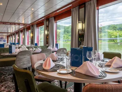 Restaurant an Bord der Belvedere: Gedeckte Tische mit blauen Menükarten und rosa Servietten. Große Fenster bieten Blick auf Fluss und bewaldete Ufer.