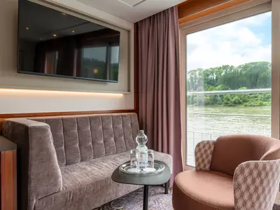 Moderne Kabine der Belvedere: Sofa, Sessel, Beistelltisch mit Getränken. Panoramafenster mit Blick auf Fluss und grüne Ufer. TV über dem Sofa.