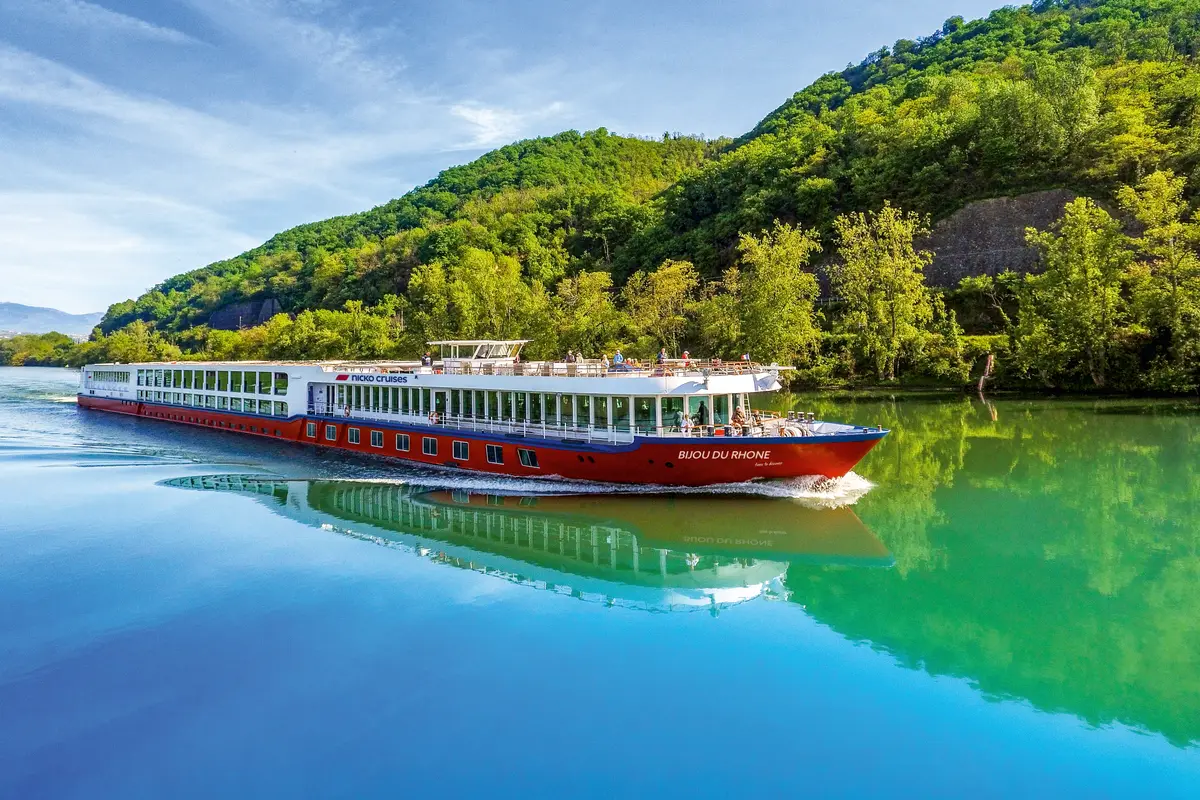 Rotes Flussschiff "Bijou du Rhone" gleitet auf einem Fluss, gesäumt von grünen Hügeln. Passagiere sind an Deck. Eine klare Spiegelung im Wasser.