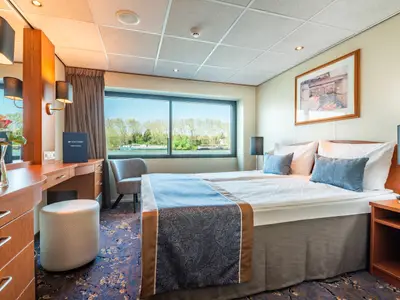 Helle Kabine an Bord der Bijou du Rhône mit Doppelbett, Schreibtisch und großem Fenster mit Blick auf Flusslandschaft und weitere Schiffe.