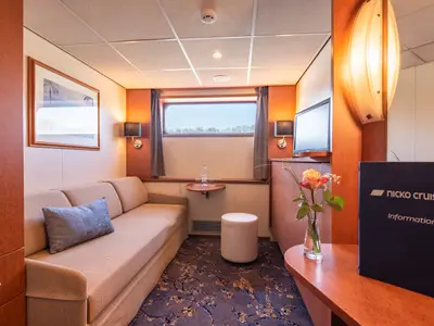 Kabine des flusskreuzfahrtschiffs bijou du rhône mit beigem Sofa, Fenster, Fernseher, Blumen und nicko cruises Informationsordner.